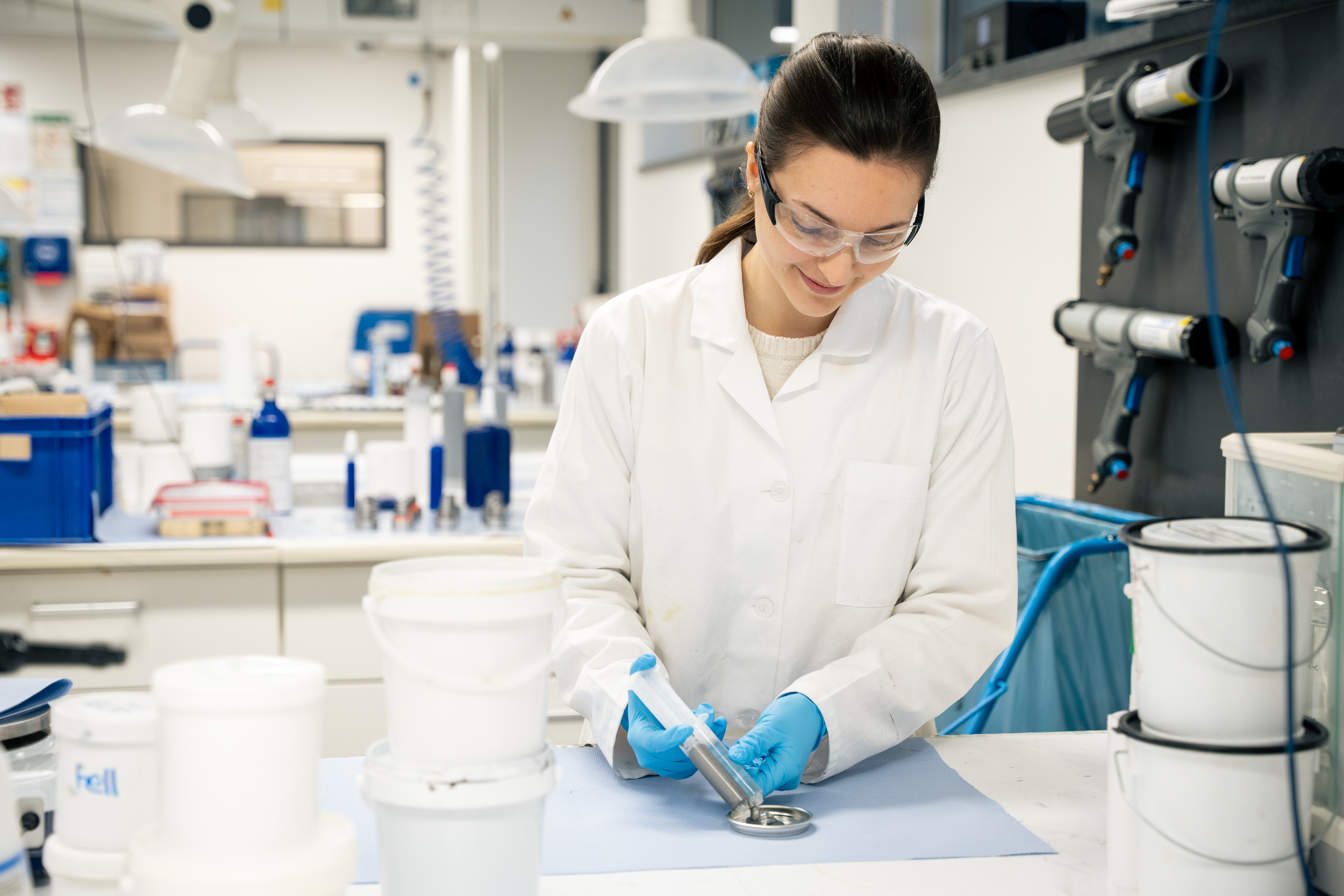 A woman wearing a white coat and safety goggles standing in a laboratory and gluing something