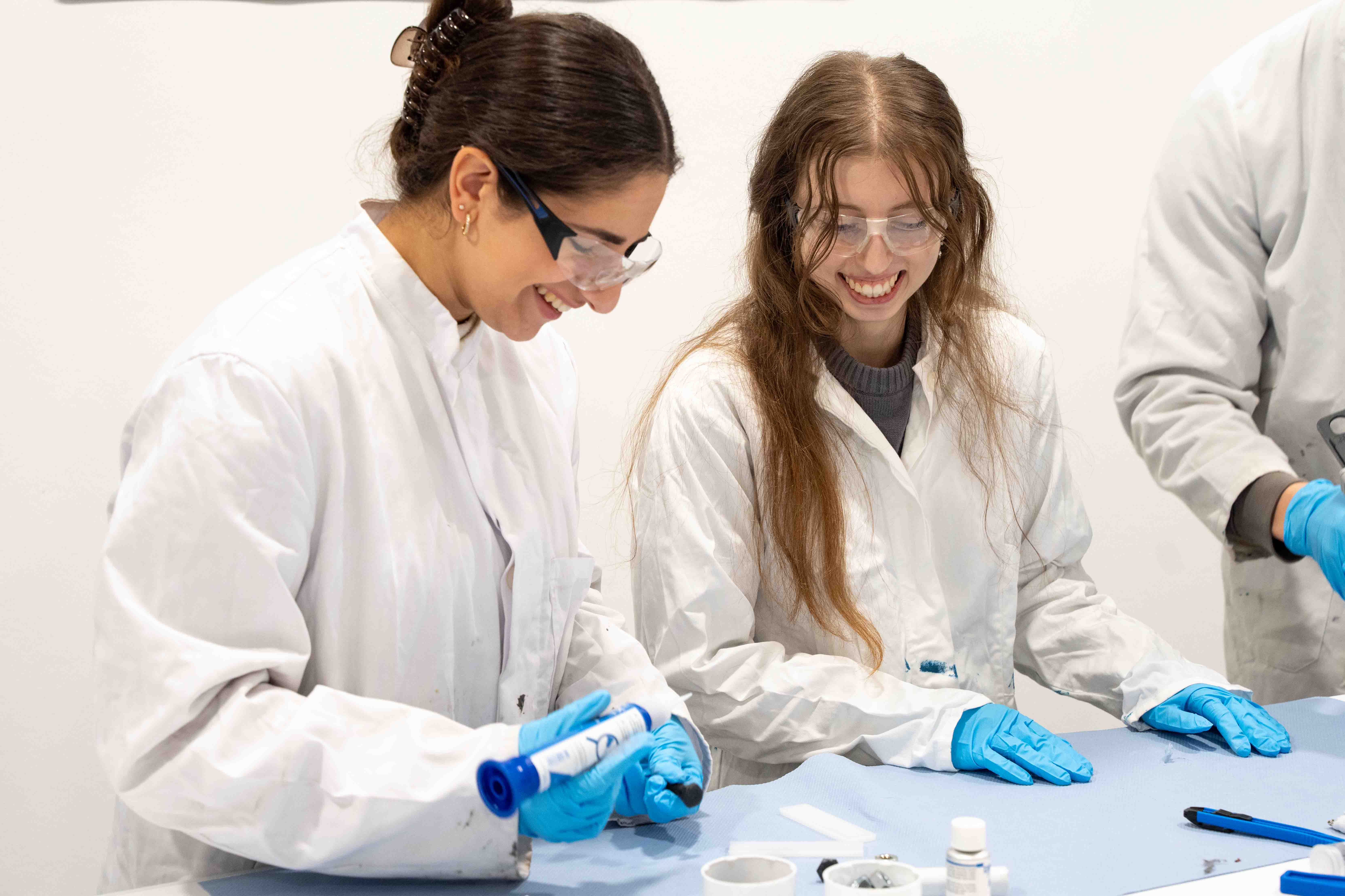 Young women in white lab coats stand laughing in a row, holding products in their hands.