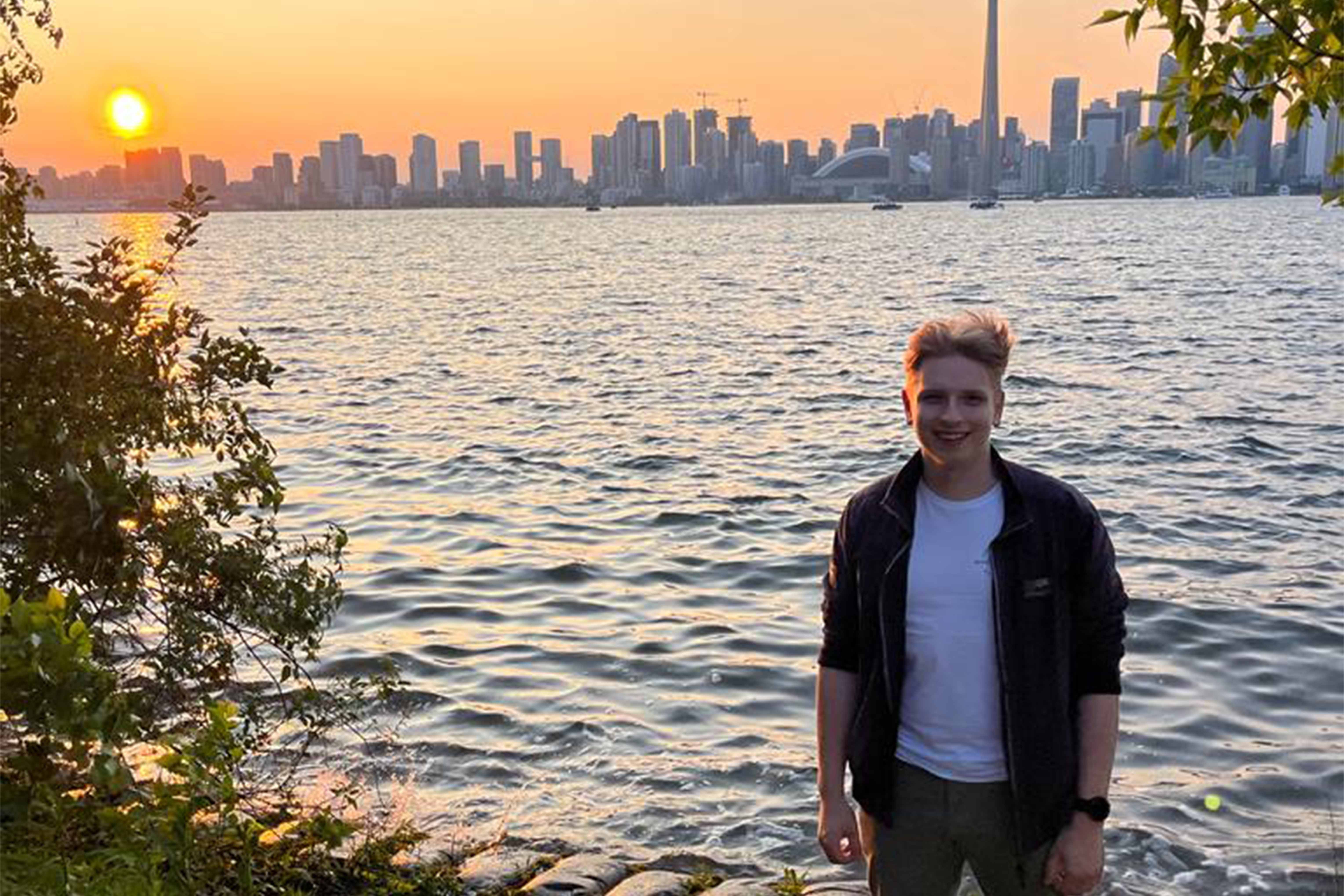 Young man standing in front of a backdrop with water, sunset and a city in the background.